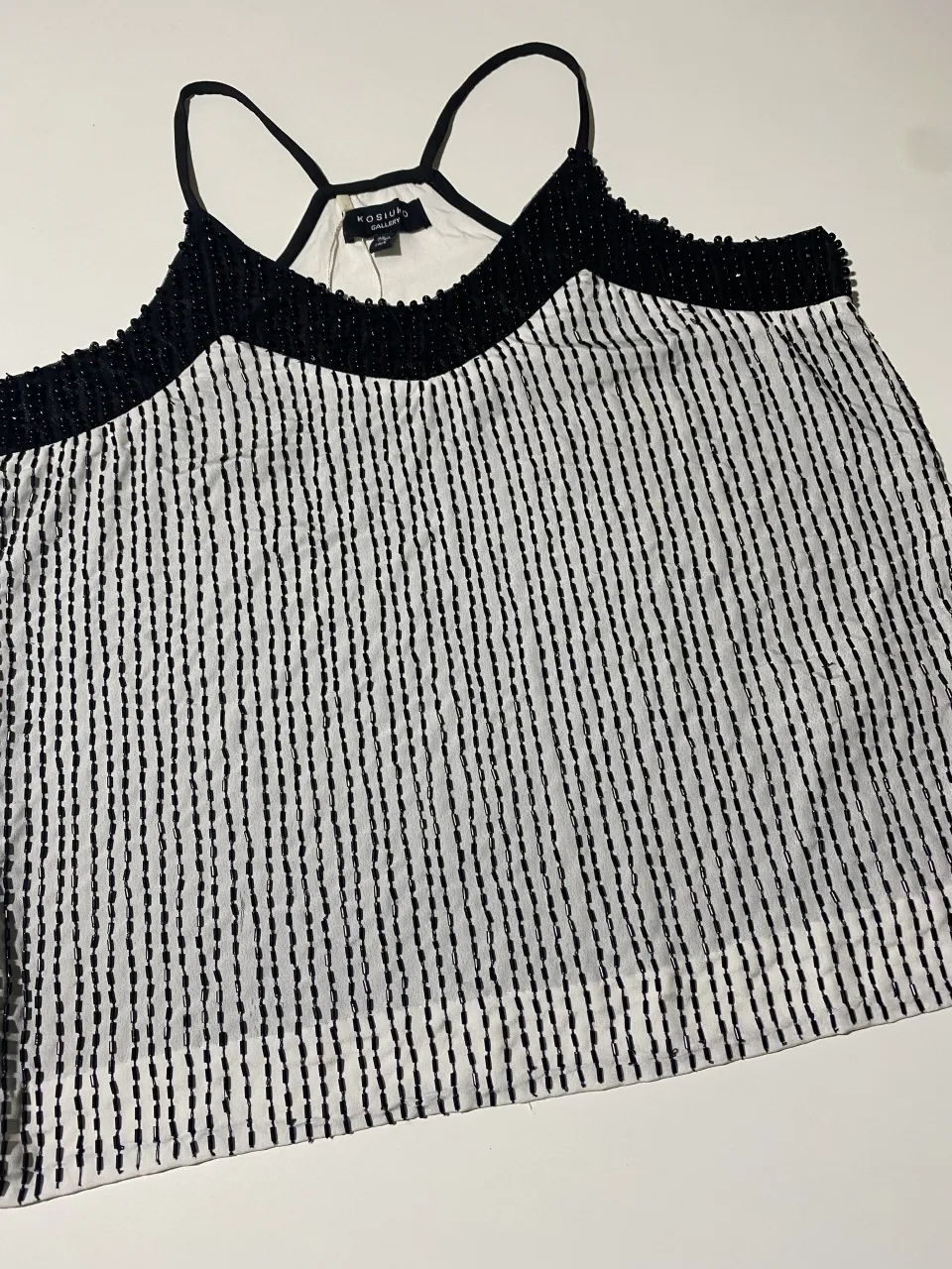 Top blanco con detalles de canutillos negros en el escote y en las tiras. Posee un estampado de finas líneas verticales negras que le dan un toque moderno y elegante. Ideal para lucir en una salida nocturna o una ocasión especial.