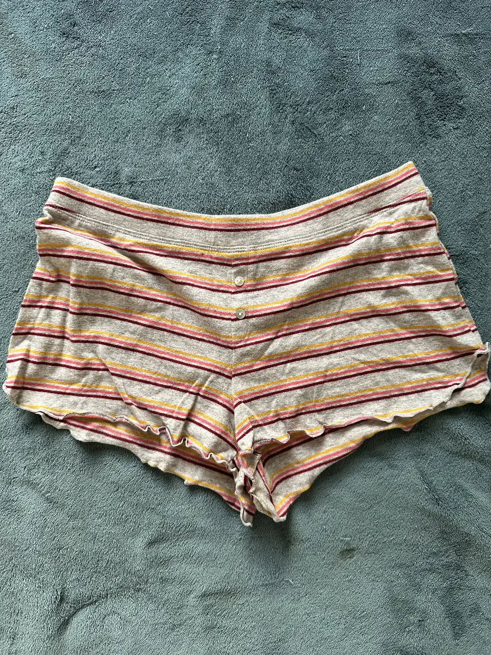Shorts pijama de tela rayada en tonos grises, rosas y amarillos, con un diseño cómodo y detalles de volados en los bordes. Ideal para un look casual y fresco.