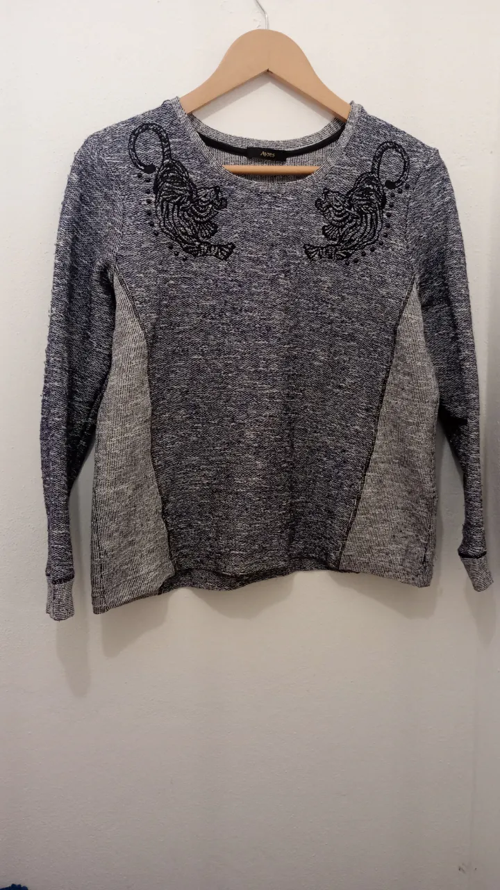 buzo tipo sweter color gris azulado gaspeado con detalles bordados de tigres en la parte delantera. Posee recortes de tela en los laterales y puños y cuello de ribb. su color también tiene un pequeño brillo plateado muy sutil que le permite también un look de noche