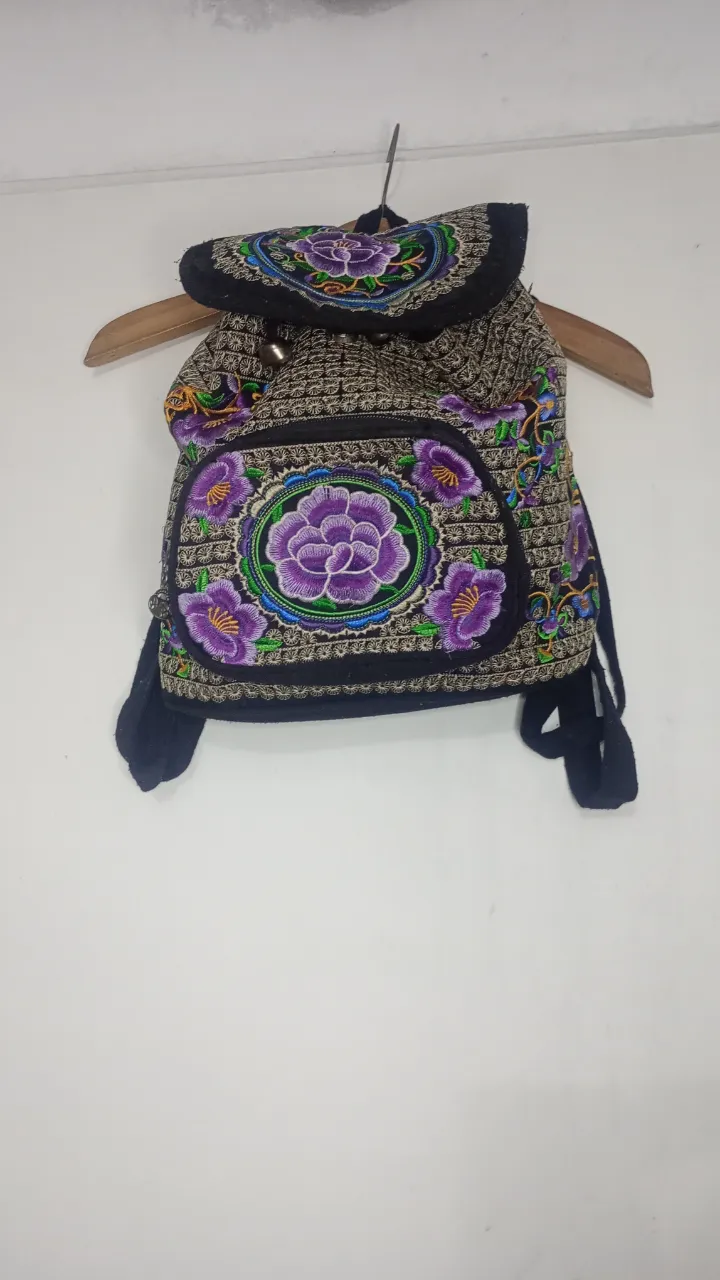 Mochila con bordados florales en tonos violeta y verde. Posee un bolsillo frontal con cierre y detalles de pasamanería. 