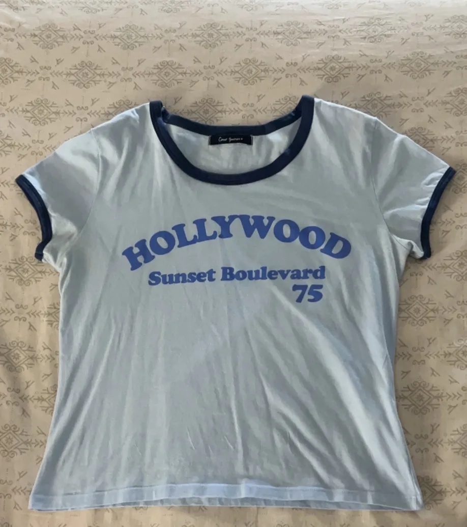 Remera manga corta color celeste con estampa "HOLLYWOOD Sunset Boulevard 75" en azul. Cuello y mangas con ribb azul marino. Ideal para un look casual y relajado. Al cuerpo.