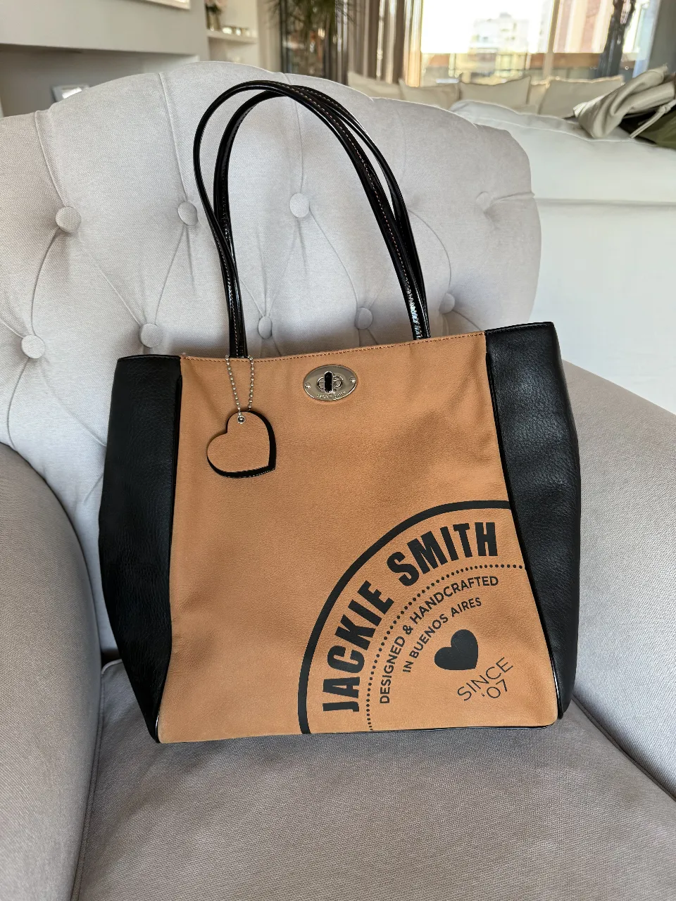 Bolso Jackie Smith de cuero sintético, con detalles en negro y marrón. Diseño con logo de la marca y dije de corazón. Ideal para llevar todo lo necesario con estilo.
