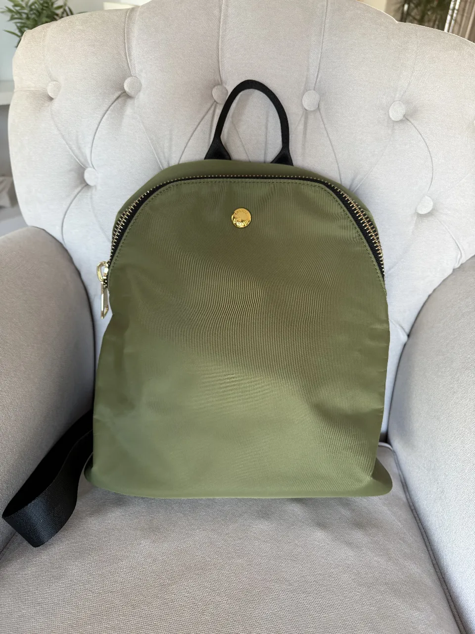 Mochila verde oliva de nailon, ideal para uso diario. Cuenta con cierre dorado, bolsillo exterior y correas ajustables. Interior negro con etiqueta de la marca.