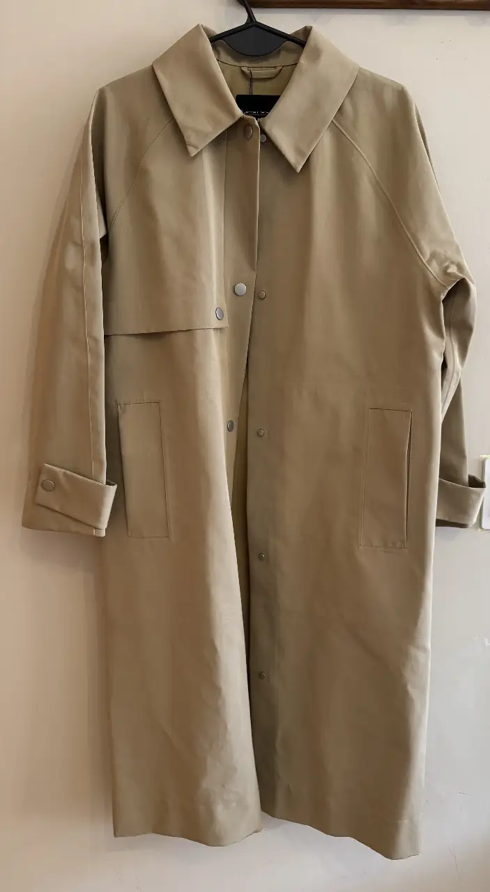 Trench coat impermeable largo de color khaki, con cuello solapa y con botones. Posee bolsillos delanteros y mangas largas con puños abotonados. Ideal para un look elegante y sofisticado.