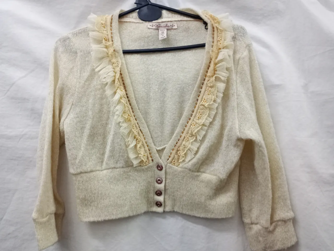 Cardigan de hilo fino color crudo con detalles de volados y perlitas en el escote. Posee botones al frente y un corte que realza la figura. Ideal para un look delicado y femenino.