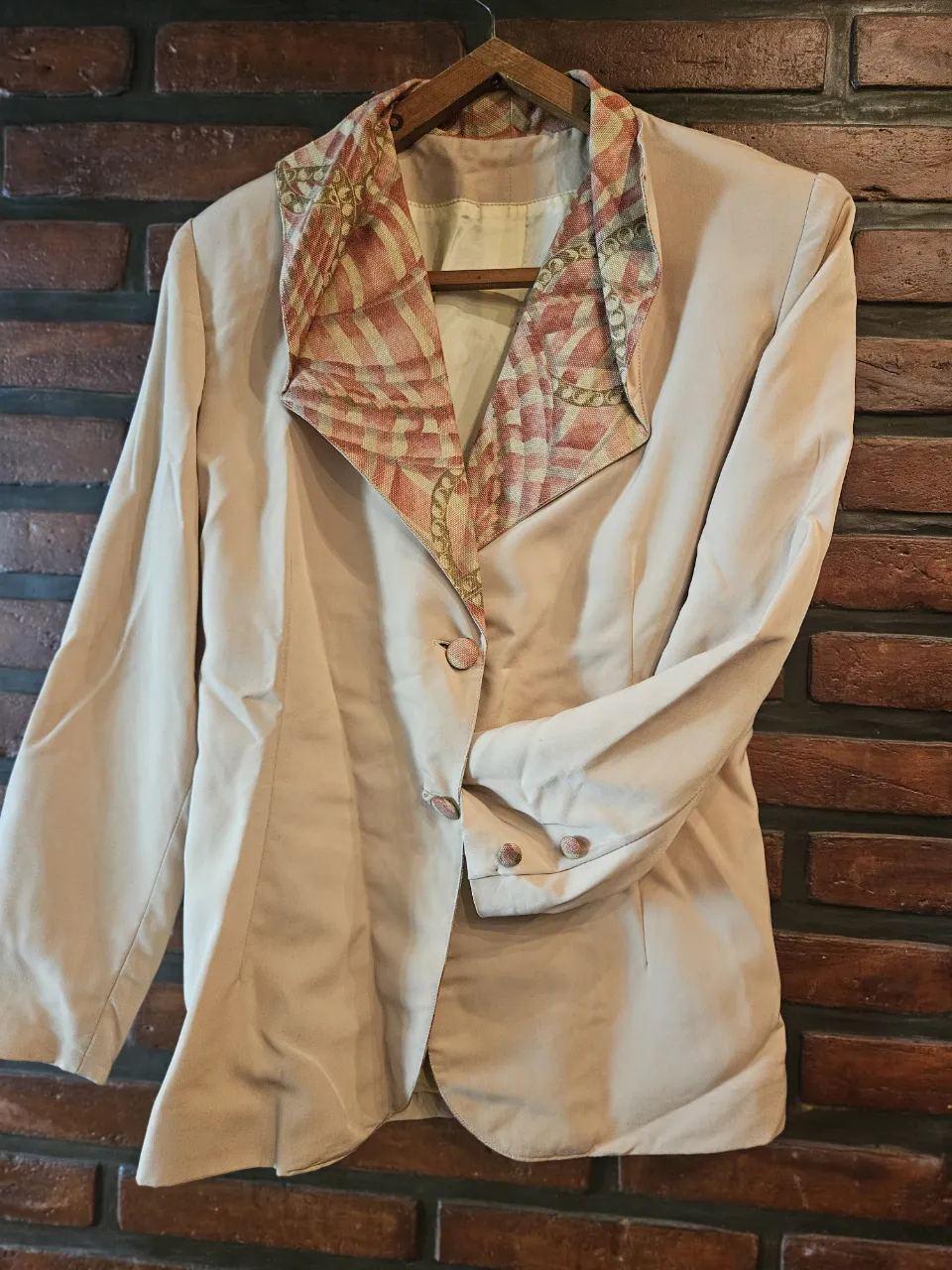 Blazer color tiza con solapa estampada en tonos rosas y dorados con diseño de cadenas. Posee un botón al frente y dos botones en cada puño. En la espalda, detalle de fruncido con hebilla plateada.