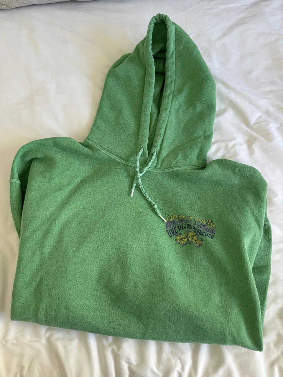 Buzo con capucha verde vibrante, con estampa "Wildest Flowers Live in San Francisco" en el frente y un diseño artístico detallado en la espalda con una mujer y flores. Ideal para un look casual y relajado.