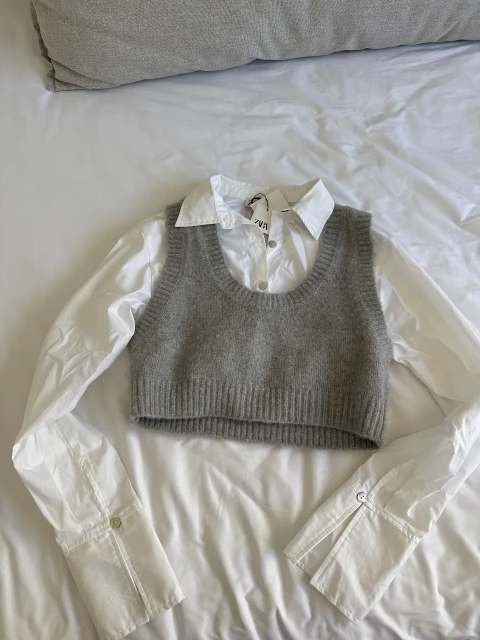Conjunto de camisa blanca de manga larga con un sweater chaleco gris de tejido de punto. El sweater es corto y tiene un escote redondo. La camisa tiene cuello y puños con botones. Ideal para un look canchero y moderno.
