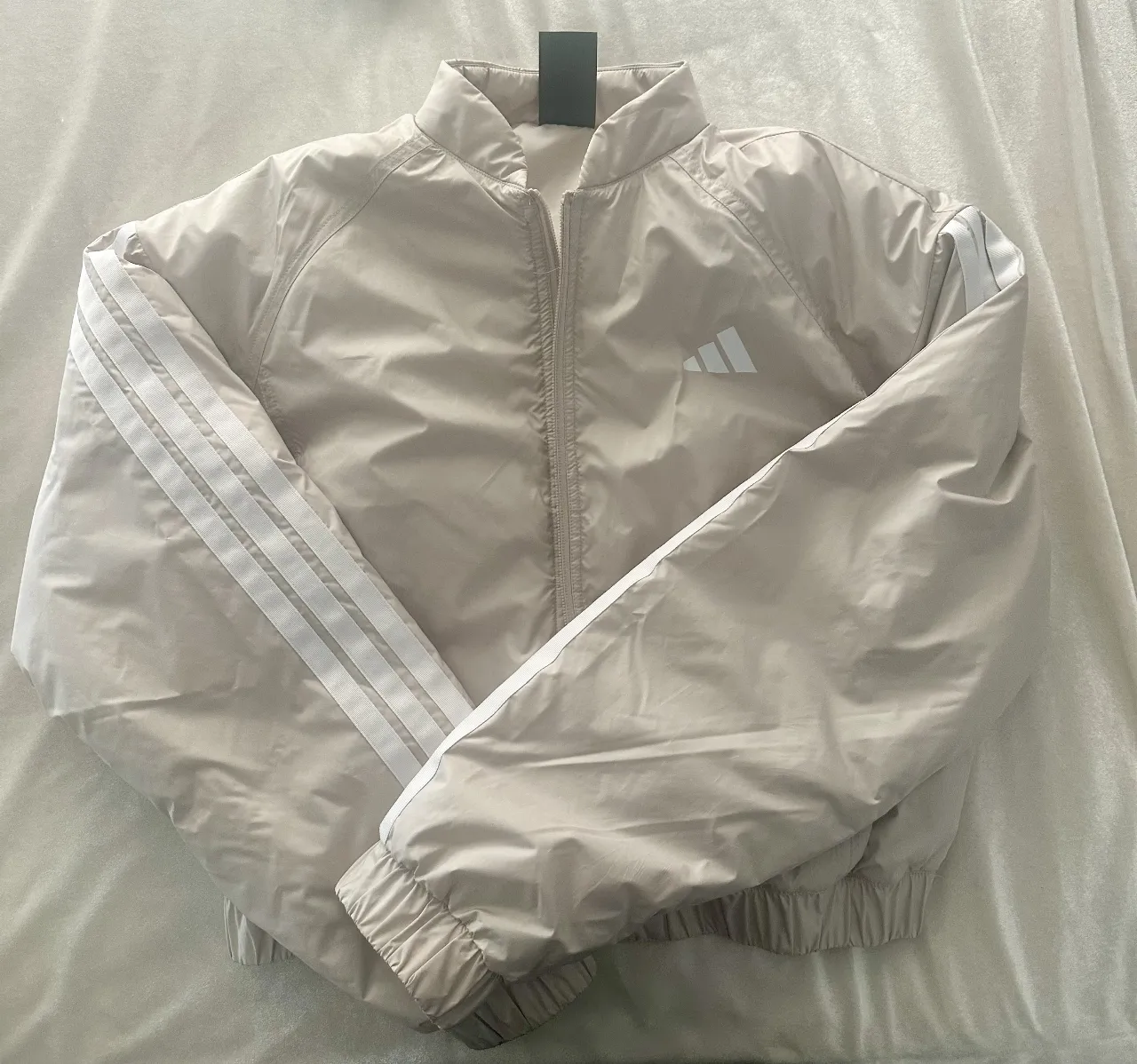 Campera bomber Adidas para mujer, color beige con las tres tiras blancas en la manga y el logo de la marca en el pecho. Ideal para un look deportivo y casual.