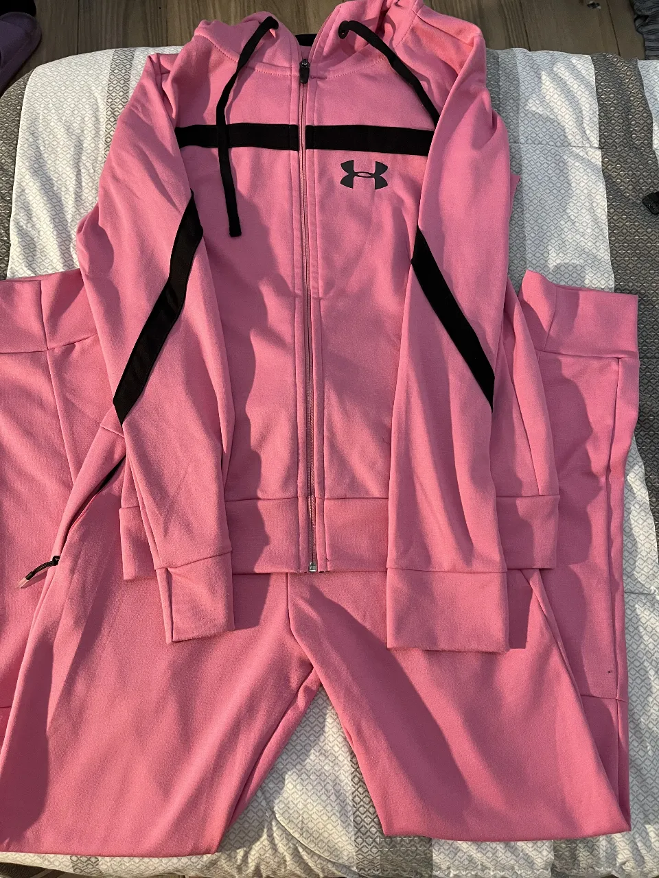 Conjunto deportivo Under Armour para mujer, color rosa con detalles en negro. Incluye buzo con cierre y capucha, y pantalón de jogging con cintura elastizada y bolsillos. Ideal para entrenar o para un look casual y cómodo.