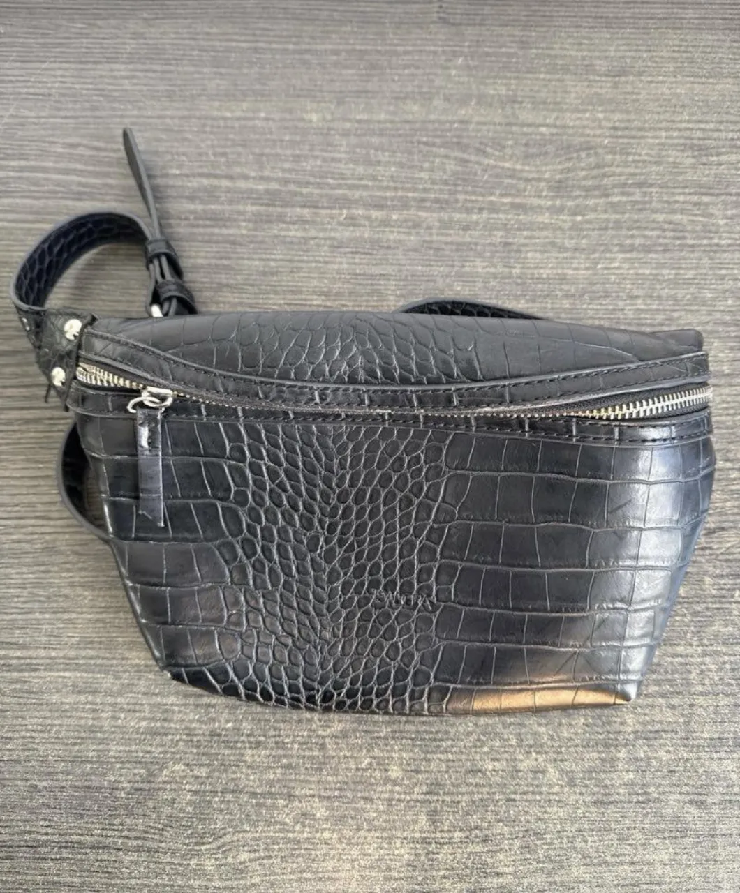 Riñonera negra de cuero con textura de cocodrilo. Tiene un bolsillo principal con cierre y una correa ajustable con hebilla.