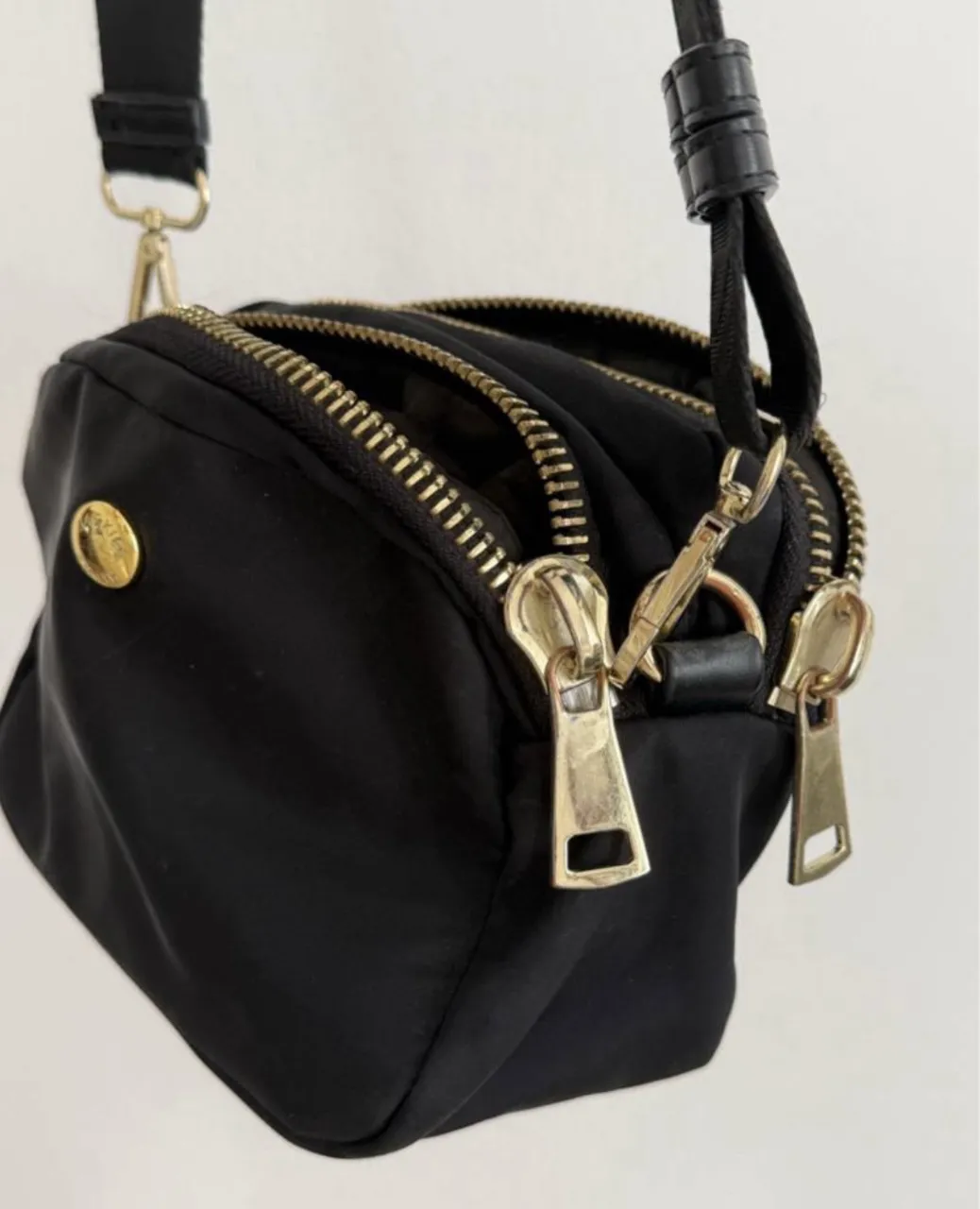 Cartera negra de nylon con tres compartimentos principales con cierre metálico dorado. Correa ajustable y logo metálico dorado en el frente. Unico detalle: desgaste en herrajes (se ve en las fotos)