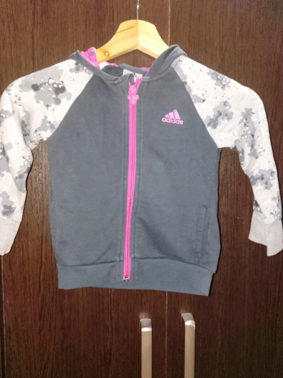 Buzo con capucha gris y mangas estampadas en camuflado blanco y gris. Posee cierre central fucsia y un aplique de Minnie Mouse en el frente. El interior de la capucha es fucsia.