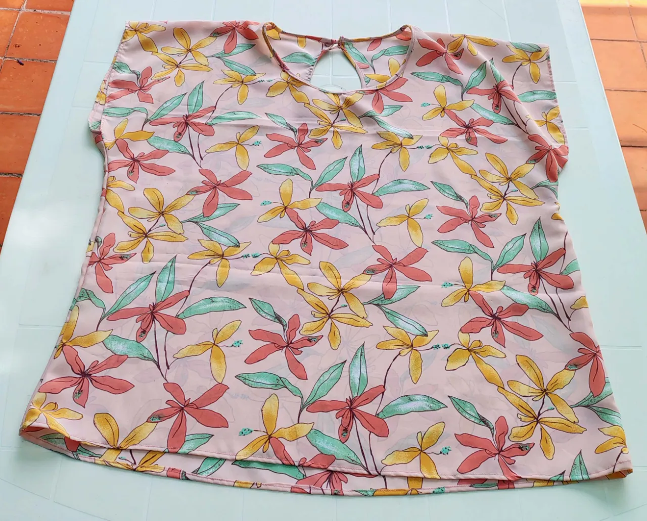 Remera de tela liviana con estampa floral en tonos pastel. Posee un recorte en forma de gota en la espalda con un botón para cerrar. Un solo uso.