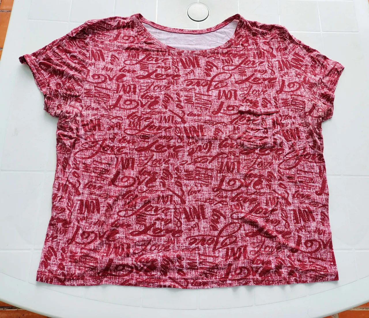 Remera de manga corta con estampado integral de la palabra "Love" en diferentes tipografías y colores rojo y blanco. Posee un bolsillo en el frente del lado izquierdo.