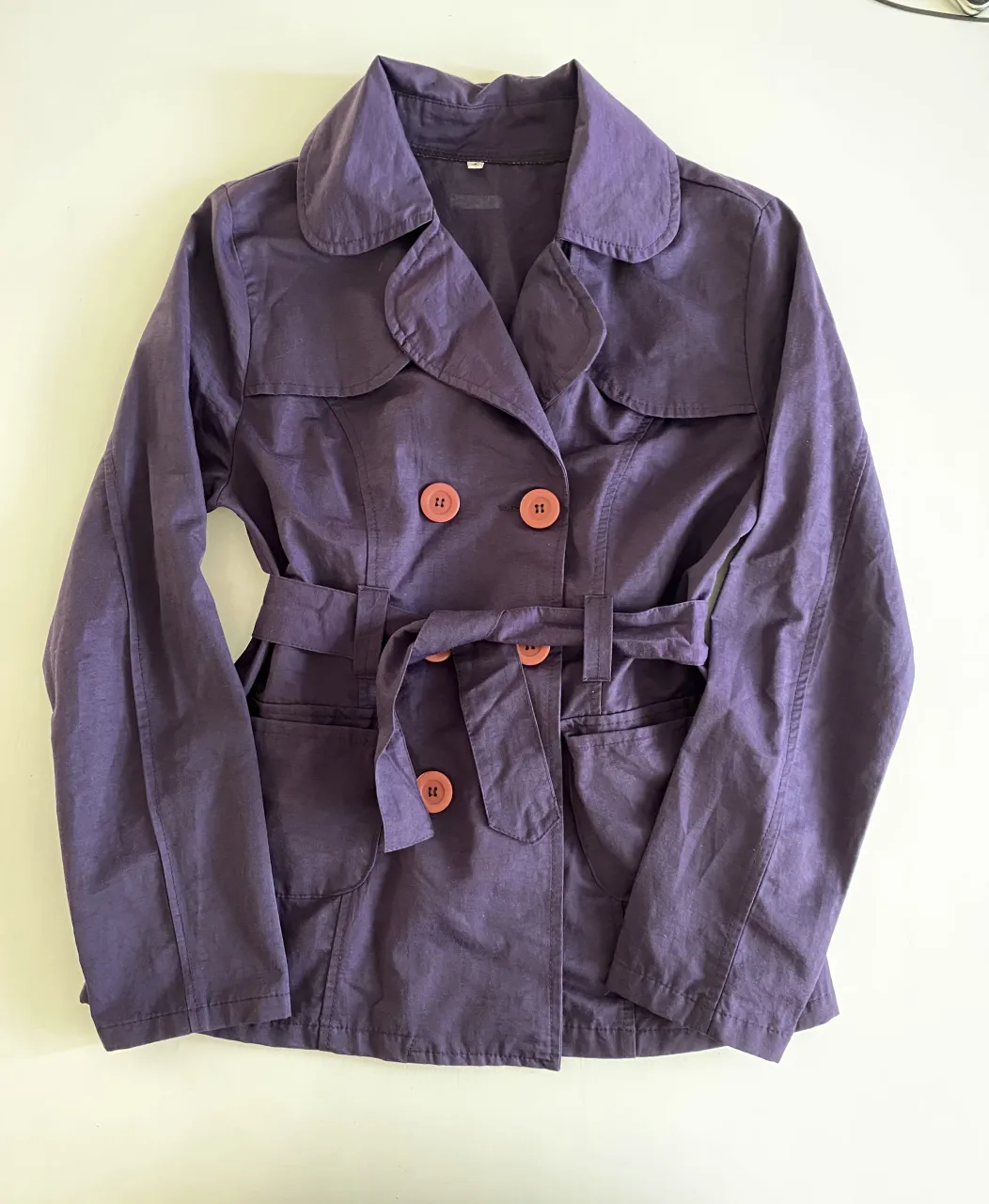 Campera corta de gabardina color violeta. Tiene cuello solapa, doble abotonadura con botones color coral, cinturón de tela para atar en la cintura y bolsillos frontales con tapa.