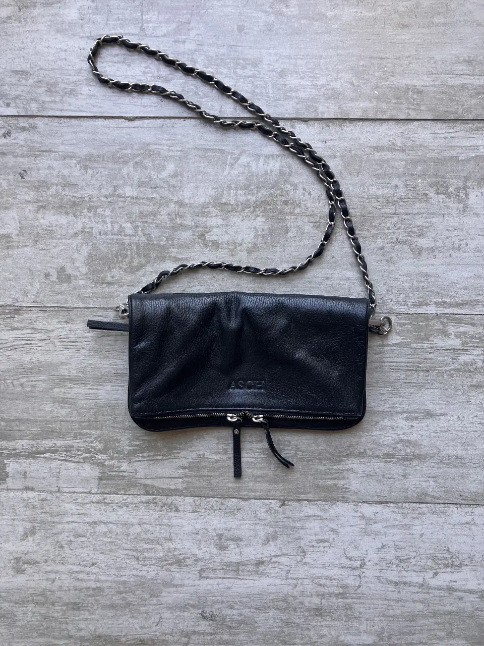 Cartera bandolera negra de cuero, con cadena plateada y cierre con detalle de flecos. Ideal para un look casual y moderno.