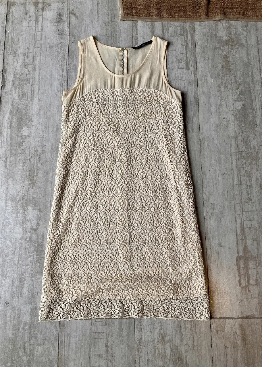 Vestido mini de mujer, color crudo. Confeccionado con una parte superior de tela lisa y una sección inferior de encaje intrincado. Cuenta con cierre en la espalda y un diseño sin mangas. Ideal para ocasiones especiales o eventos formales.