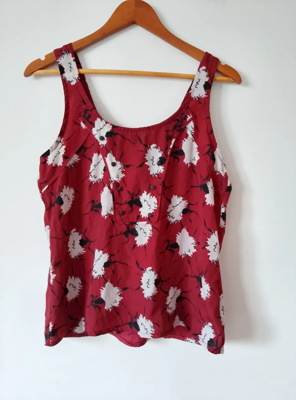 Blusa sin mangas de color rojo con estampado floral blanco y negro. Tiene botones decorativos en la parte delantera y un corte clásico.