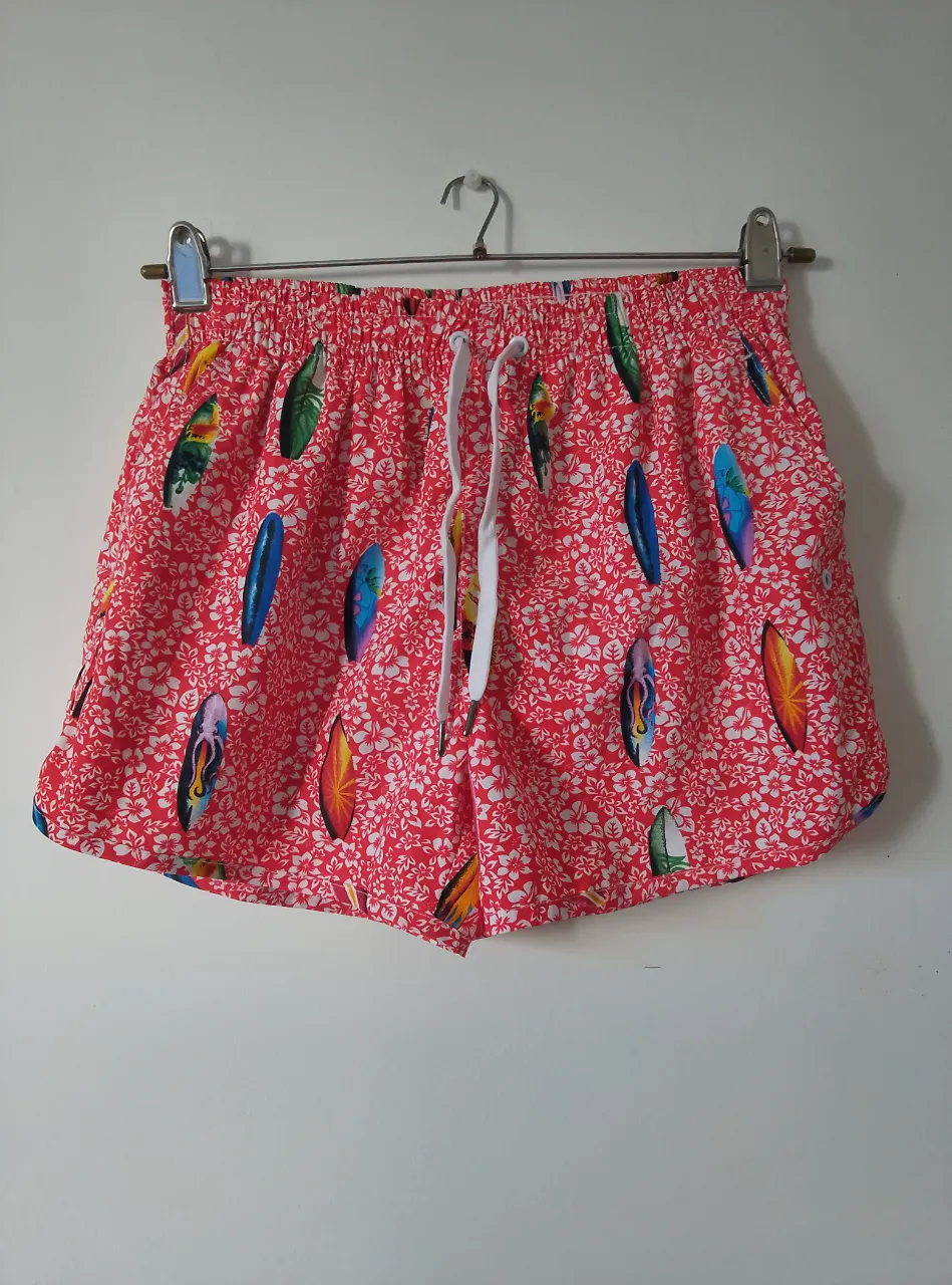 Short de baño para hombre, con estampado de tablas de surf sobre fondo rojo y flores blancas. Cuenta con cordón blanco en la cintura y forro interior de malla.