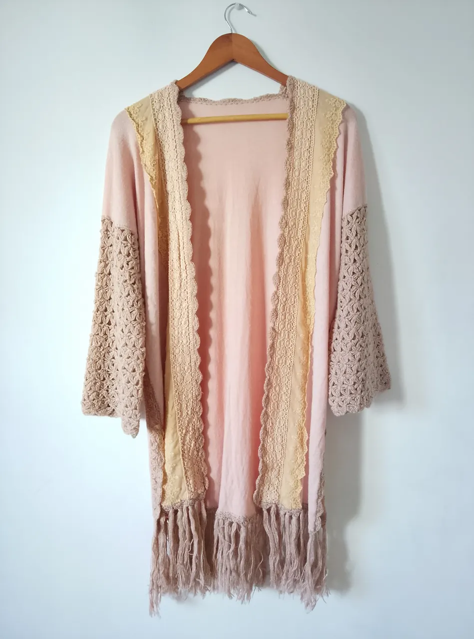 Kimono cardigan de tejido liviano en color rosa con detalles en beige y flecos. Mangas amplias con detalles calados. Ideal para un look bohemio y relajado.