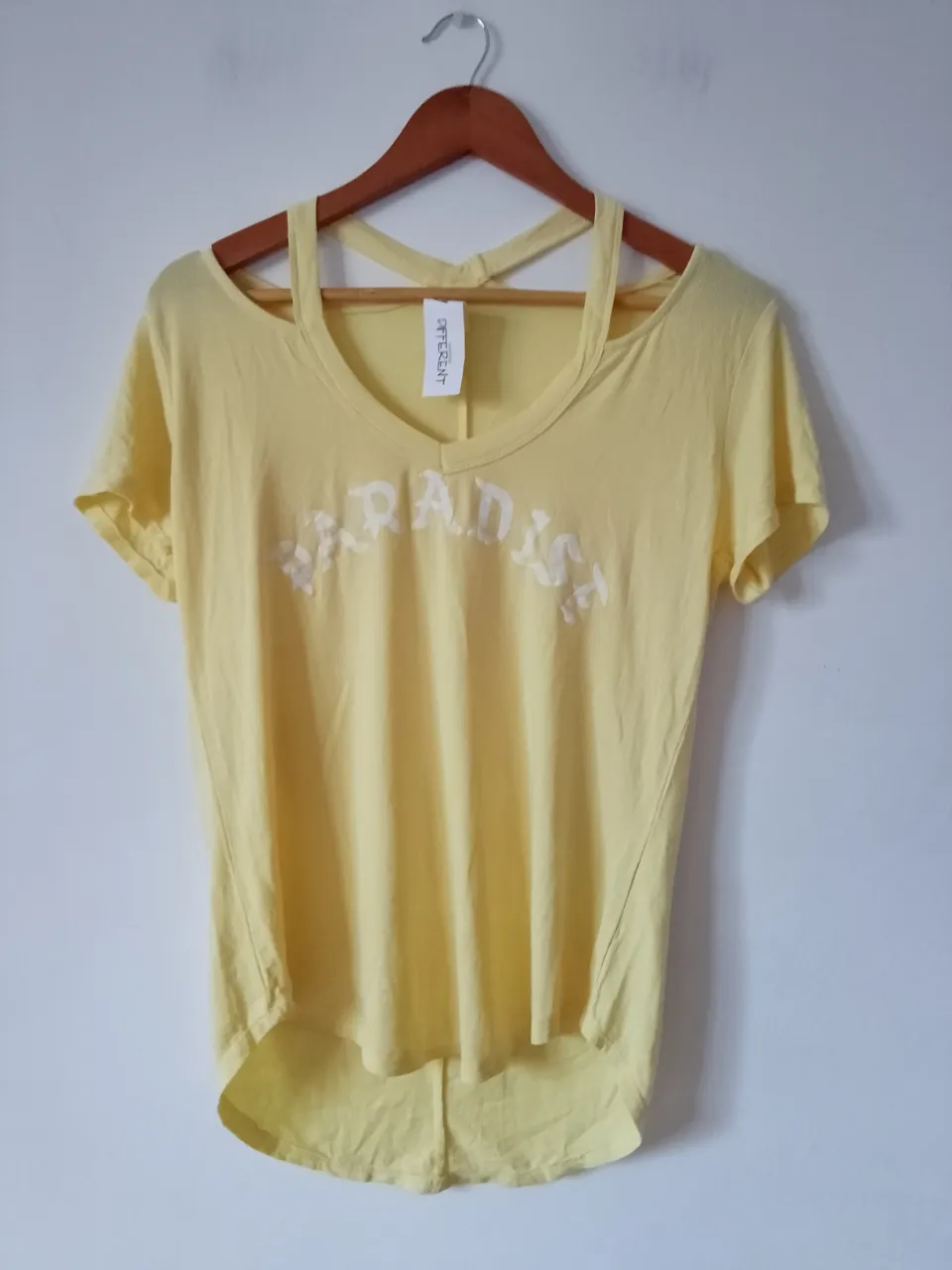 Remera amarilla con estampado "PARADISE" en blanco. Diseño con hombros descubiertos y corte asimétrico. Ideal para un look casual y fresco.