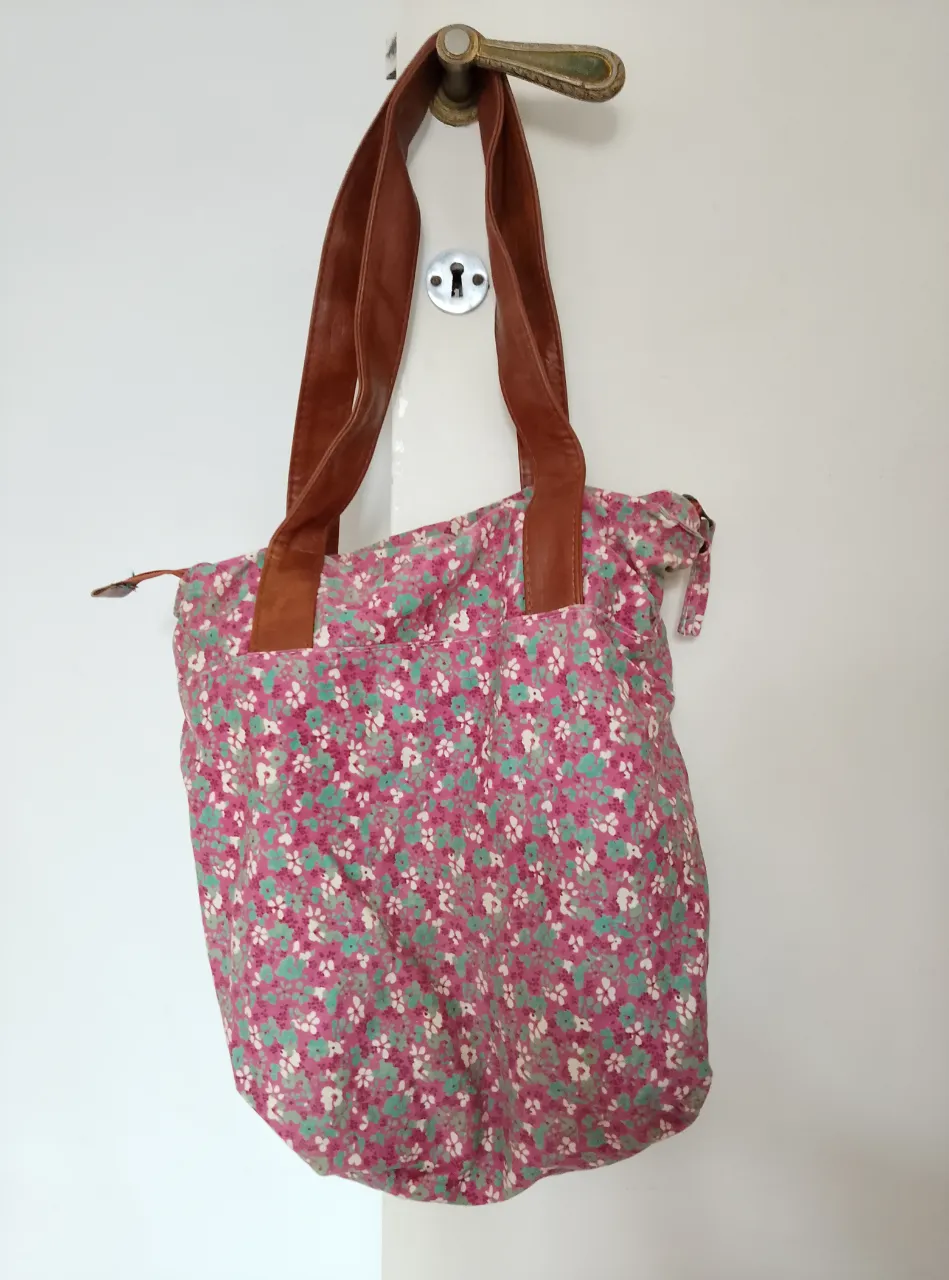Bolso de playa de tela floreada en tonos rosa y verde, con manijas de cuero marrón. Ideal para llevar todo lo necesario con estilo.