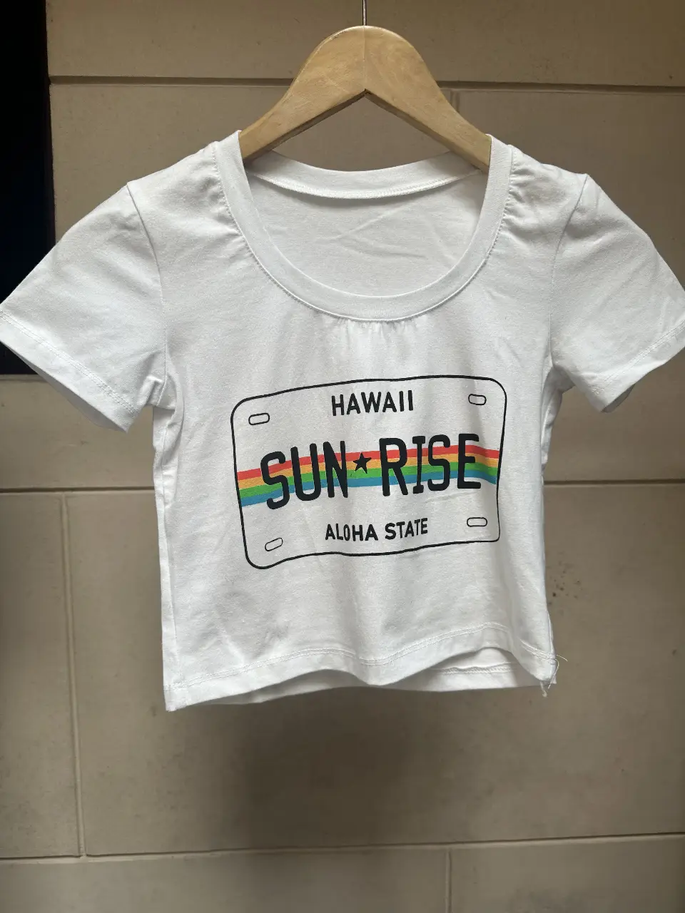 Remera crop top blanca con estampado de matrícula de Hawaii. Diseño con arcoíris y la frase 'SUN RISE'. Ideal para un look casual y playero. talle único.