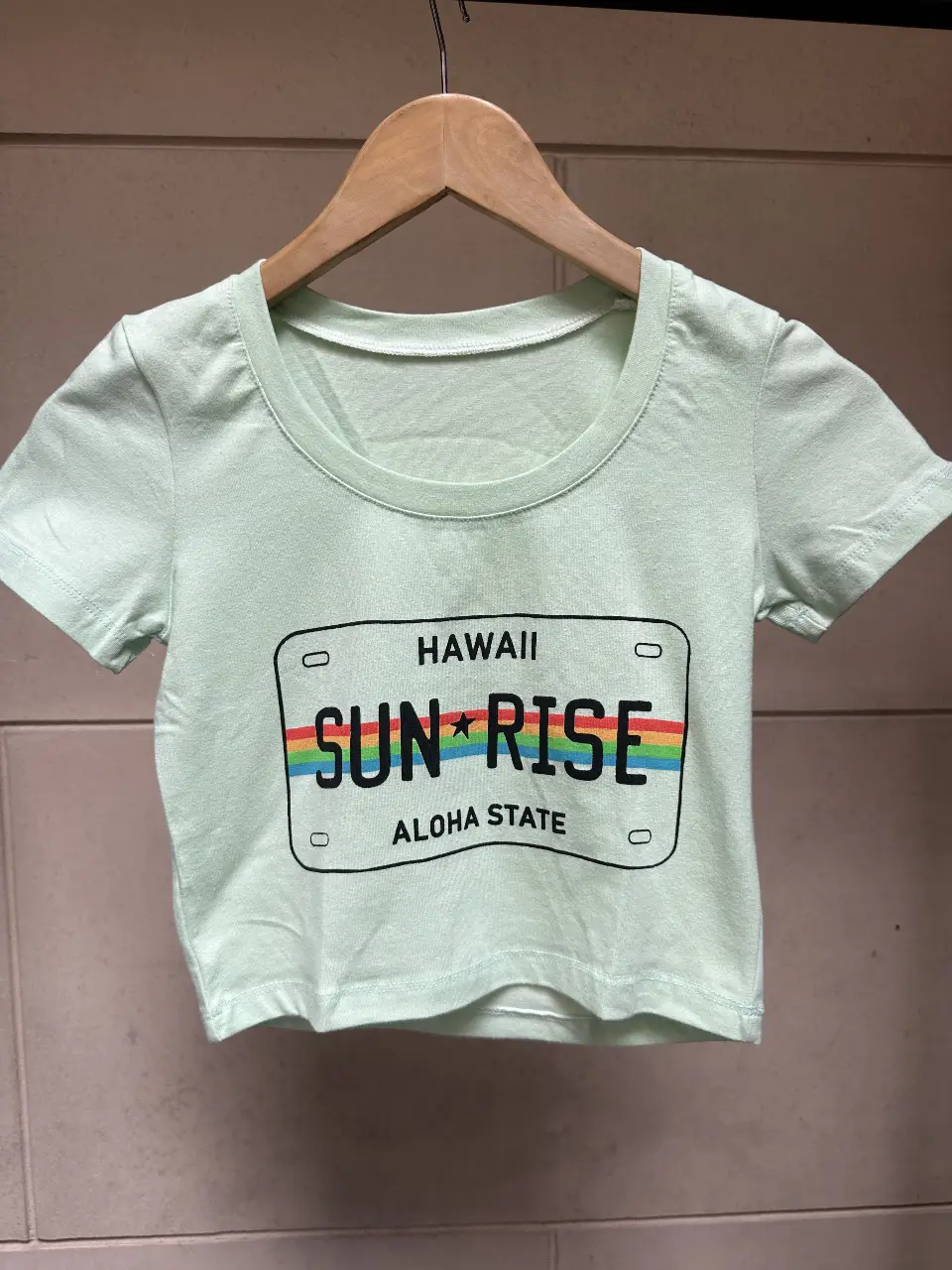 Remera crop de color verde claro con estampado de licencia de Hawaii con la palabra "SUN RISE" y un arcoíris. Ideal para un look casual y fresco. talle único 