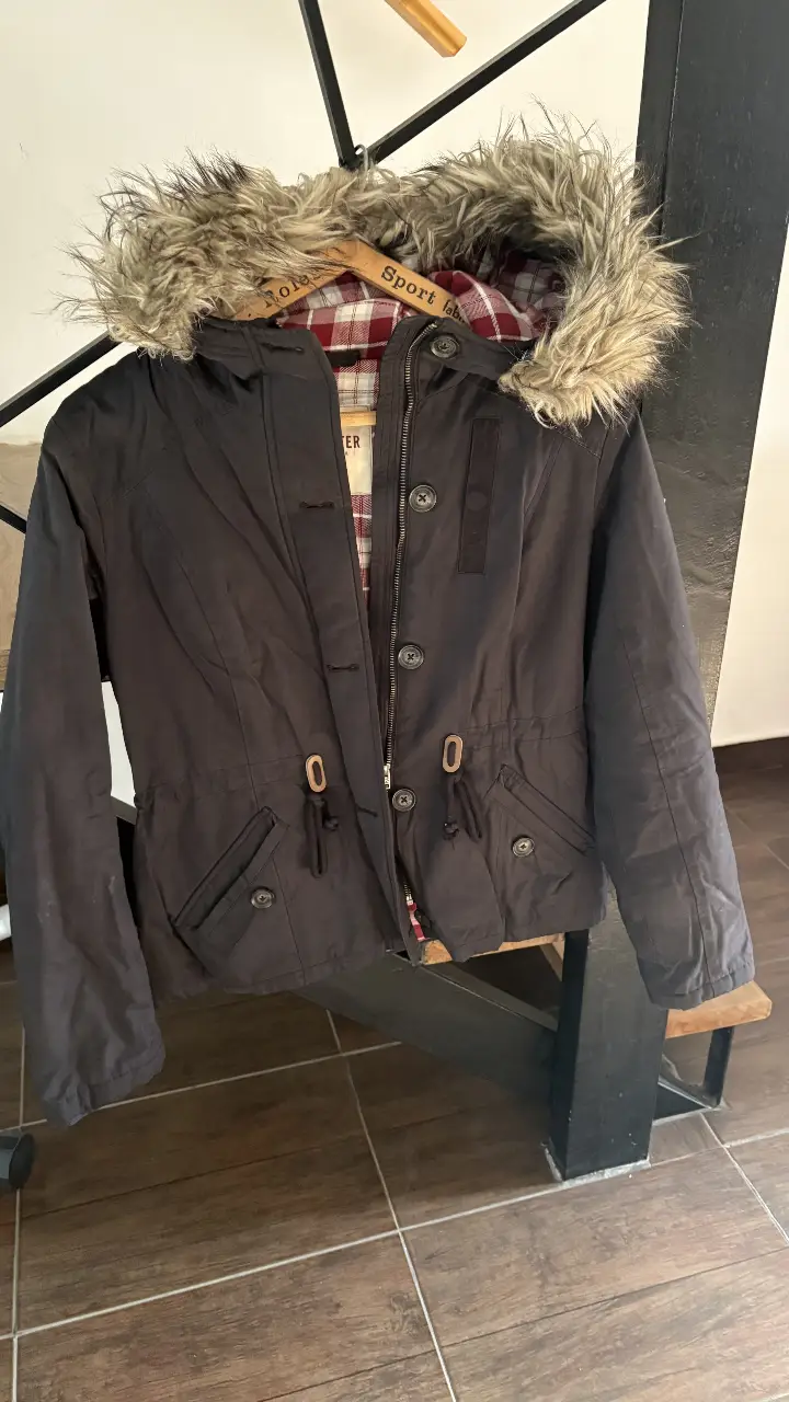 Campera Hollister negra con capucha y borde de piel sintética. Interior con forro a cuadros rojo y blanco. Talle M.