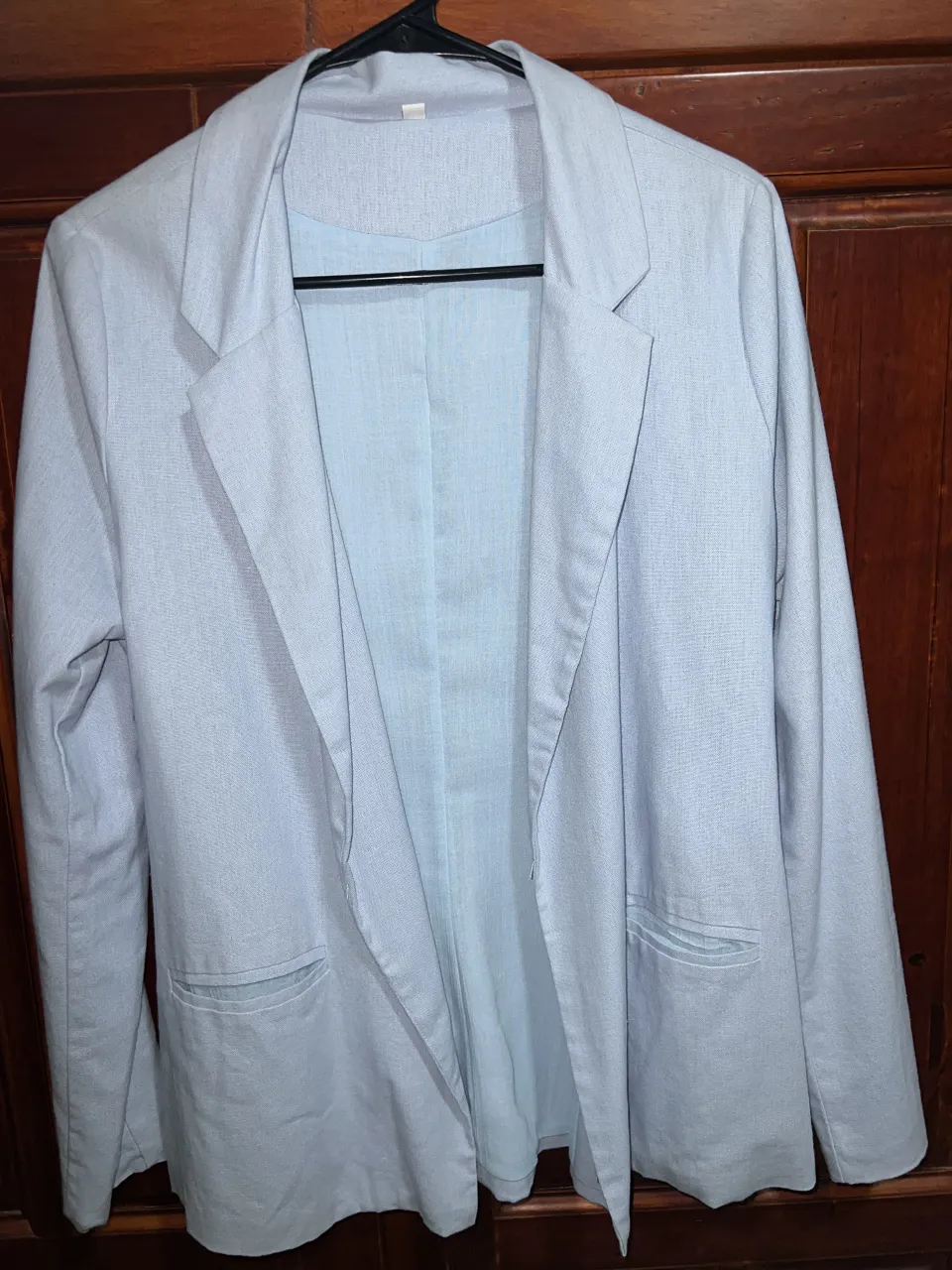 Blazer de color celeste claro, con solapas y bolsillos frontales. Se muestra colgado y también puesto sobre una remera azul. Blazer para primavera/verano. 