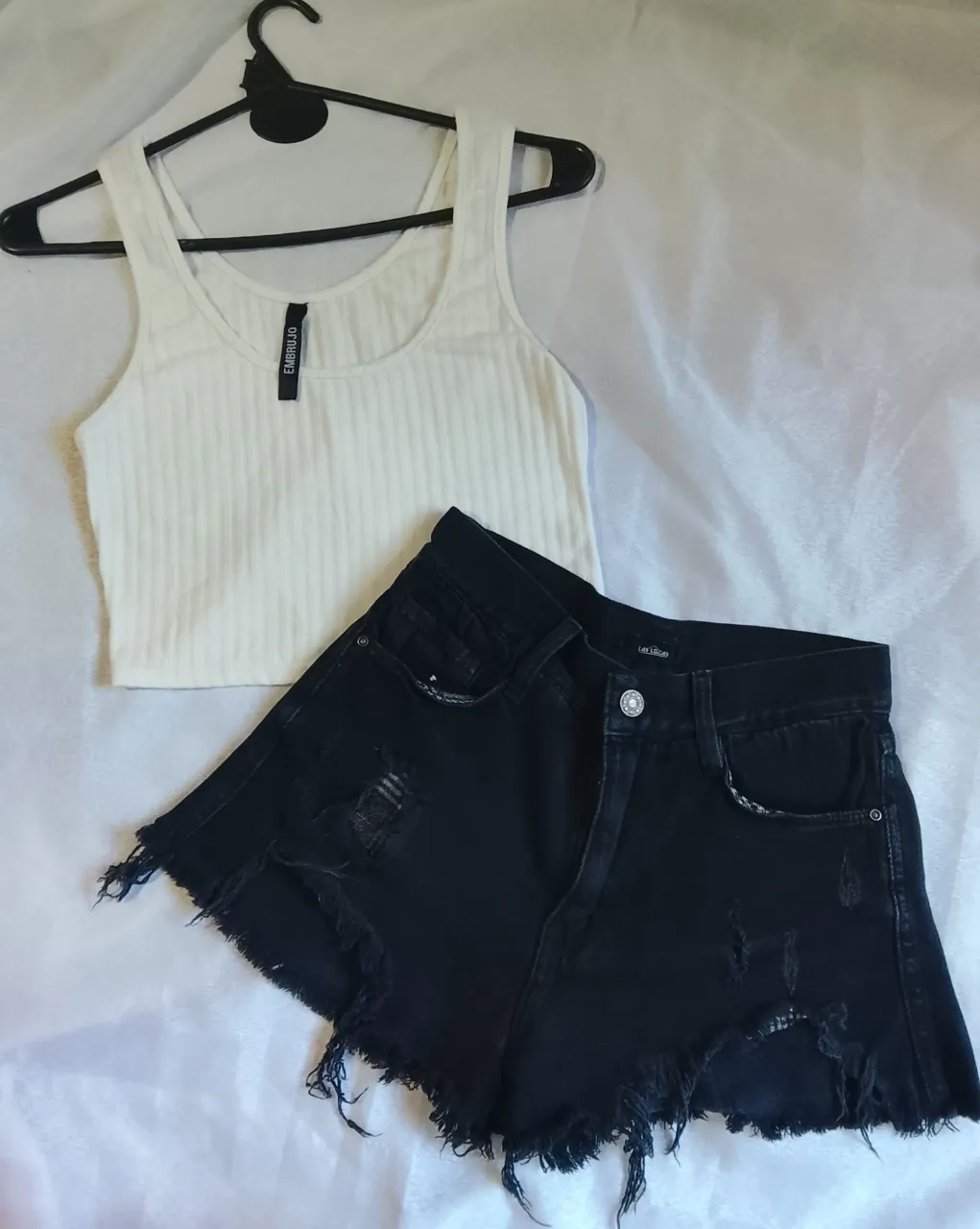 Remera blanca de morley, escote redondo y breteles anchos. Short de jean negro, tiro alto, con roturas y detalles deshilachados en los bordes.