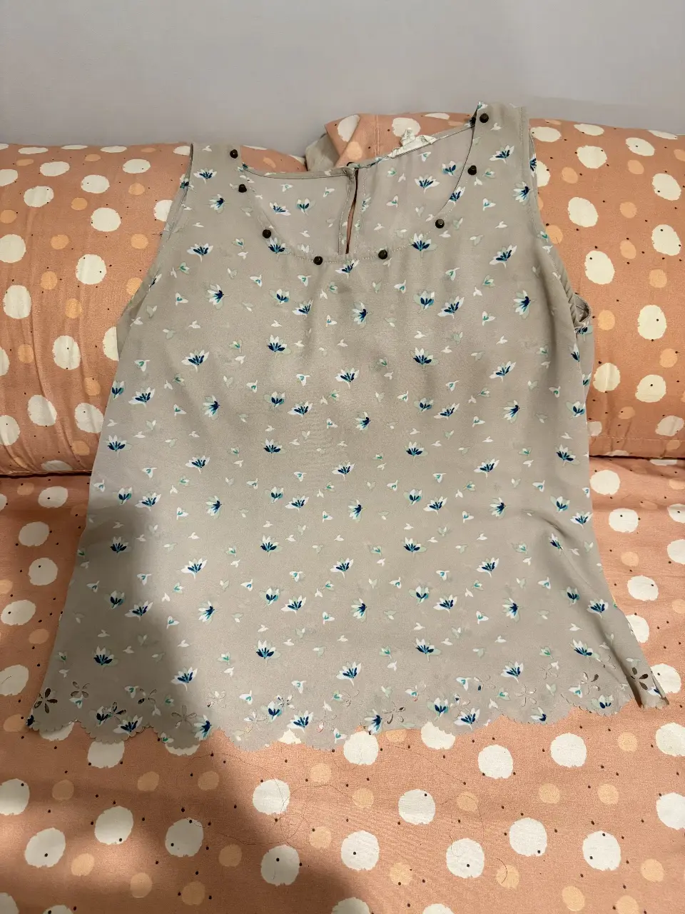 Blusa sin mangas de color gris con estampado floral en tonos azules y blancos. Tiene detalles de botones en el cuello y un borde inferior con diseño calado. Ideal para un look fresco y femenino.