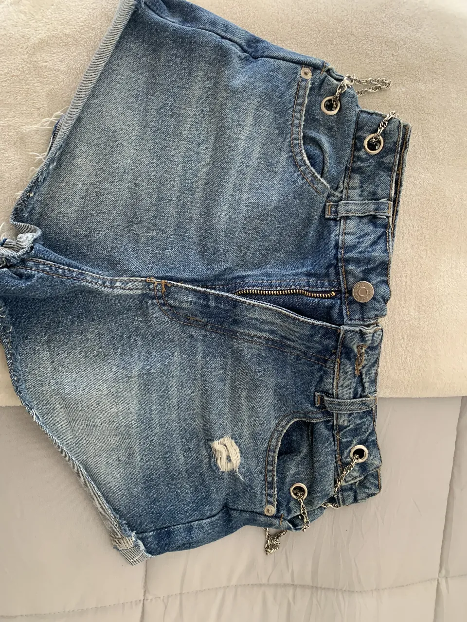 Short de jean azul con detalles de cadenas plateadas en los laterales. Tiene un corte desflecado y un pequeño roto en la parte delantera. Ideal para un look casual y moderno.