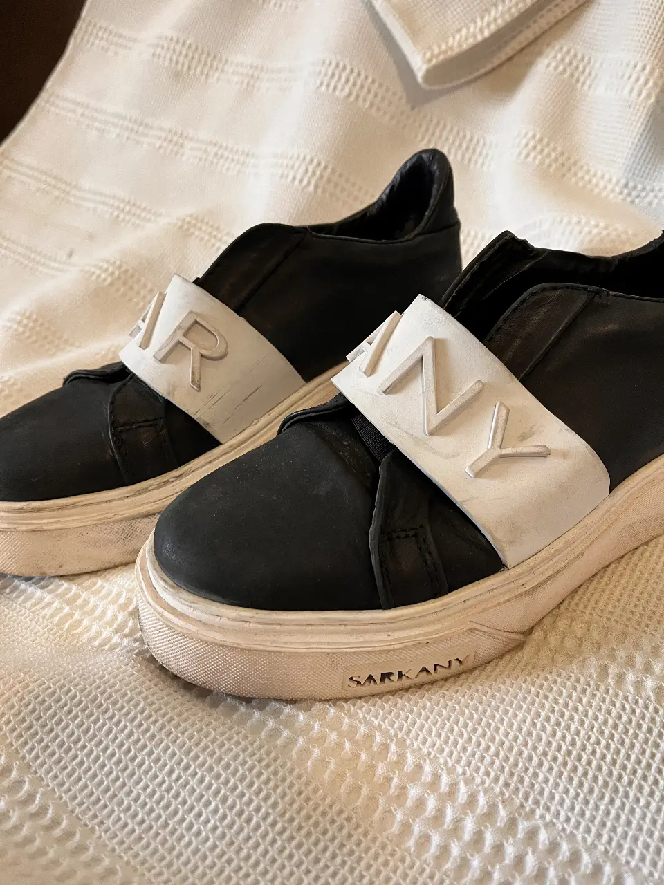 Zapatillas Sarkany negras con detalles blancos. Diseño slip-on con banda ancha en el empeine con el nombre de la marca. Suela blanca con detalles de desgaste.