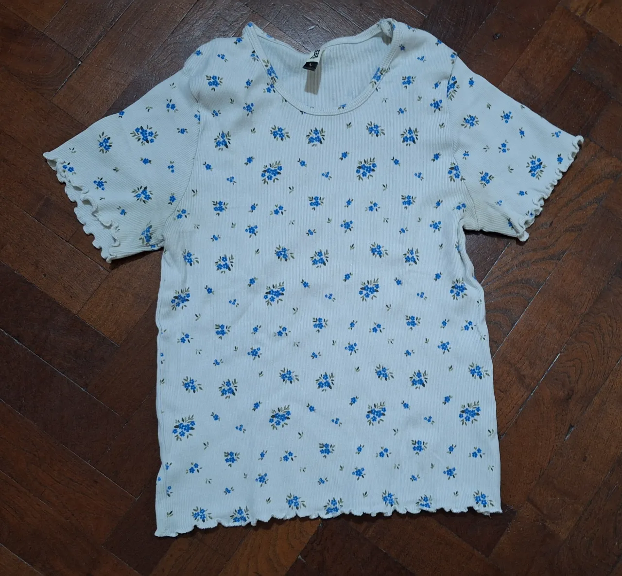 Remera blanca con estampa de flores azules y hojas verdes. Posee mangas cortas y terminaciones con volados.