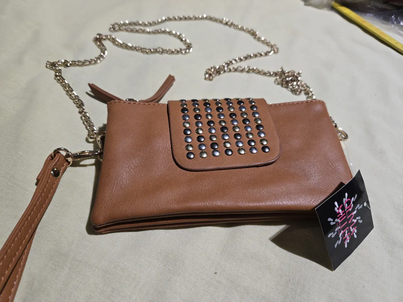 Cartera marrón con tachas doradas y plateadas en la solapa. Posee correa de cadena dorada y muñequera desmontable. El interior tiene forrería animal print.