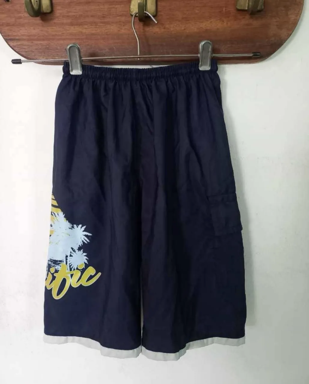 Short de baño azul marino con estampado "Pacific" en amarillo y blanco. Diseño con bolsillo lateral y detalles en blanco en el borde inferior. Ideal para la playa o actividades acuáticas.