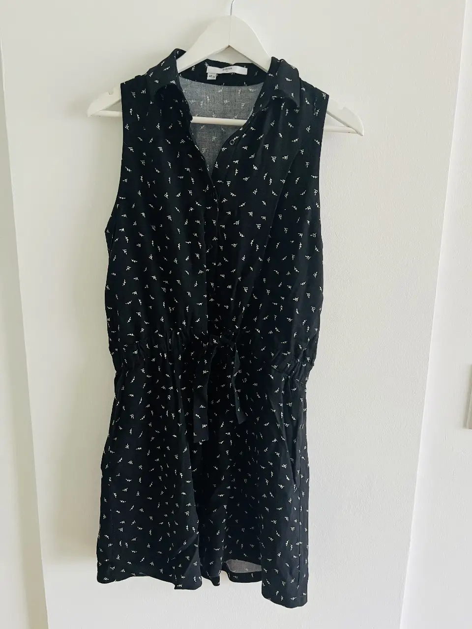 vestido corto negro con estampado de pequeños diseños blancos. Tiene cuello camisero, cierre con botones, bolsillos y un cinturón para ajustar la cintura. Ideal para un look casual y cómodo.