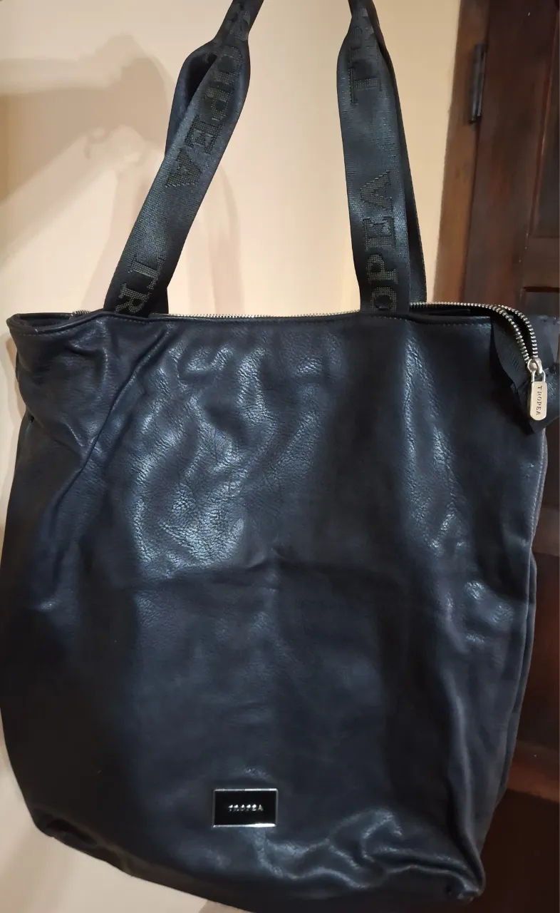 Bolso negro de cuero sintético con dos manijas de cinta negras con logo repetido en gris. Cierre superior con detalle de tirador metálico con logo.