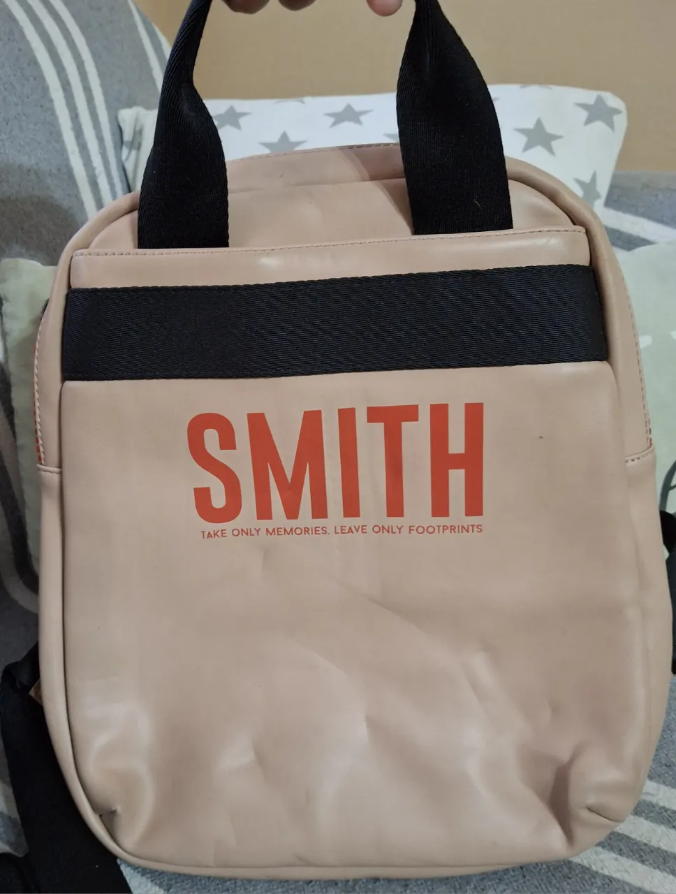 Mochila rosa pálido con la inscripción "SMITH" en color naranja y la frase "TAKE ONLY MEMORIES. LEAVE ONLY FOOTPRINTS." debajo. Posee dos tiras negras en la parte superior y una tira negra horizontal en el frente.