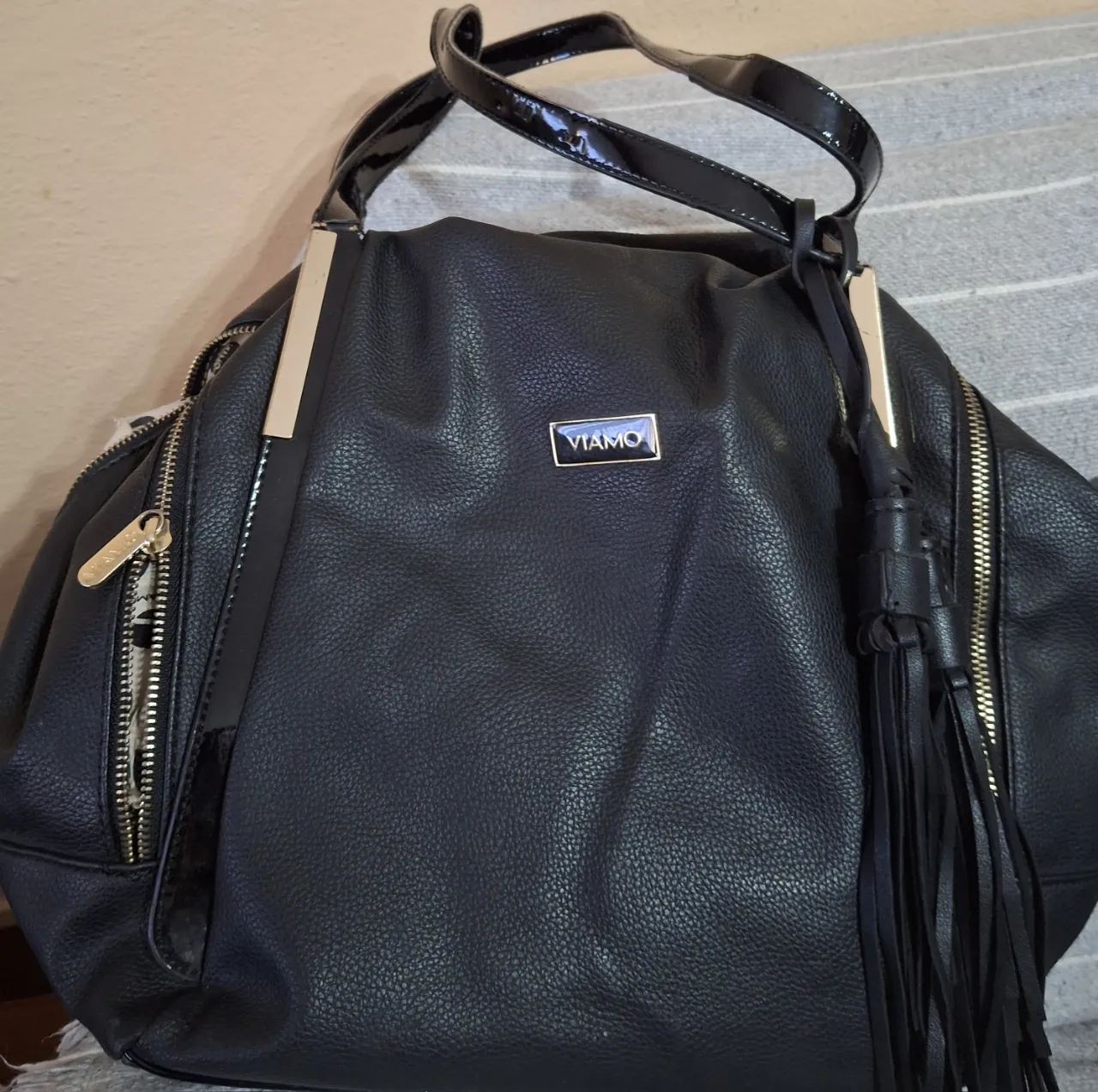 Bolso negro de cuero texturizado con dos asas cortas y una larga de charol. Tiene detalles de cierres metálicos dorados a los costados y un aplique metálico con el logo de la marca en el frente. Incluye un detalle de flecos largos colgando.