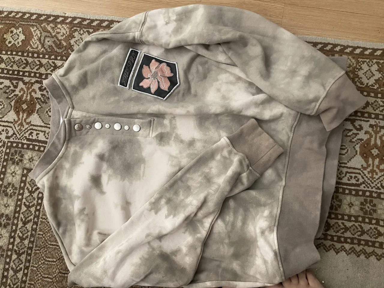 ¡Sweater oversize con estampa tie-dye súper canchero! Tiene un diseño único con detalles de botones en el cuello y un parche bordado con una flor en el frente. Perfecto para un look relajado y con onda.