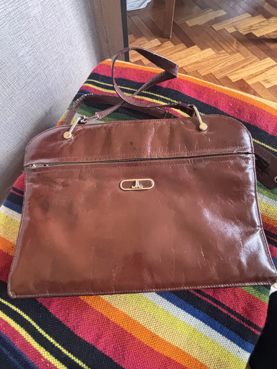 Cartera de cuero marrón, con cierre principal y un bolsillo exterior con cierre. Posee un logo metálico dorado en el frente.