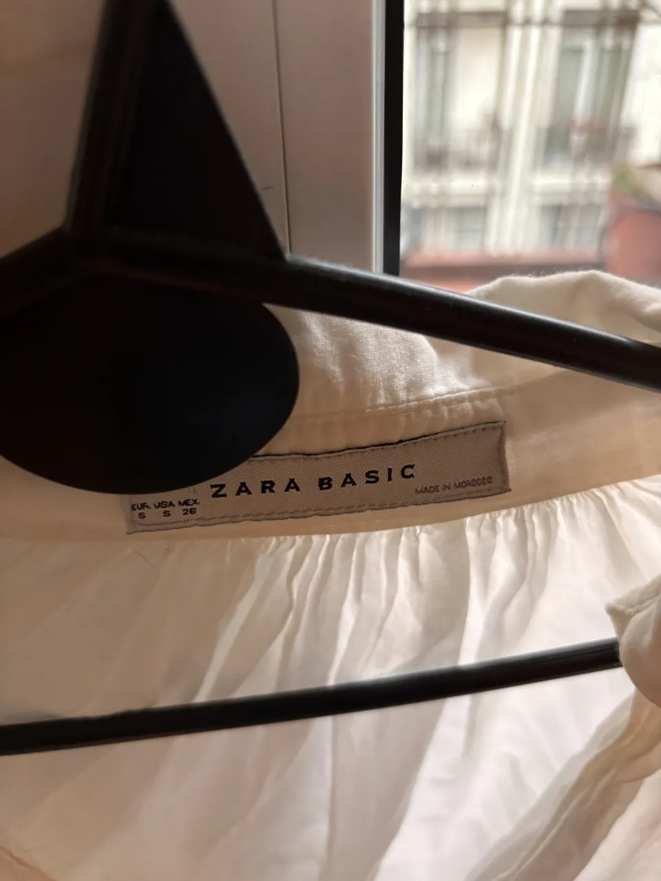 Blusa blanca de manga larga con cuello camisero y escote en V. Posee detalles de frunces en la parte superior.