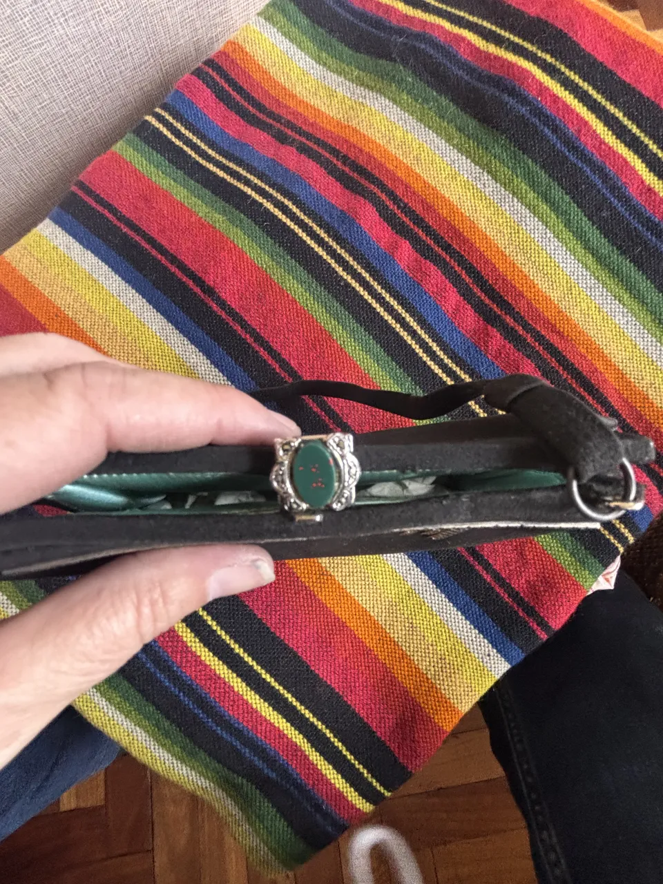 Cartera negra con detalles plateados en la parte superior y un broche central con una piedra verde. Tiene una correa de tela negra.
