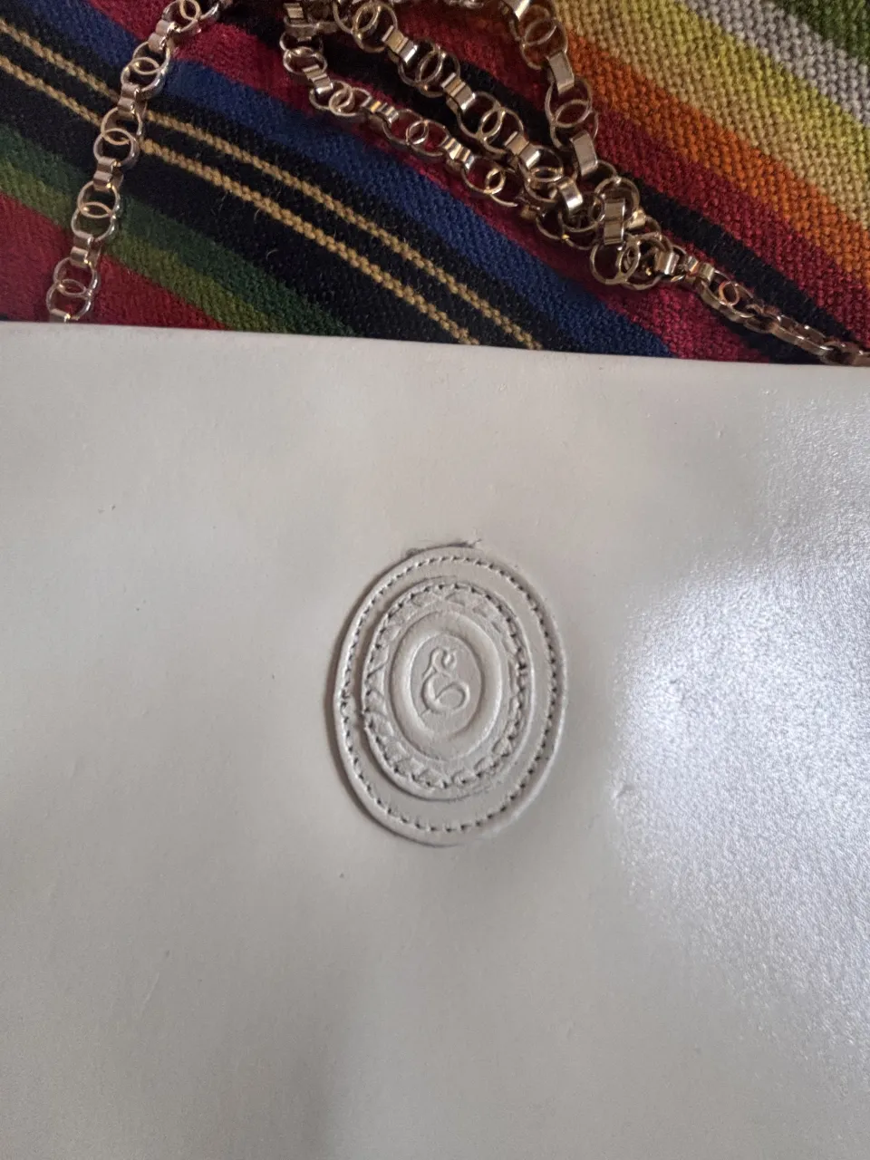 Cartera blanca con cadena dorada y logo circular en relieve en el frente.