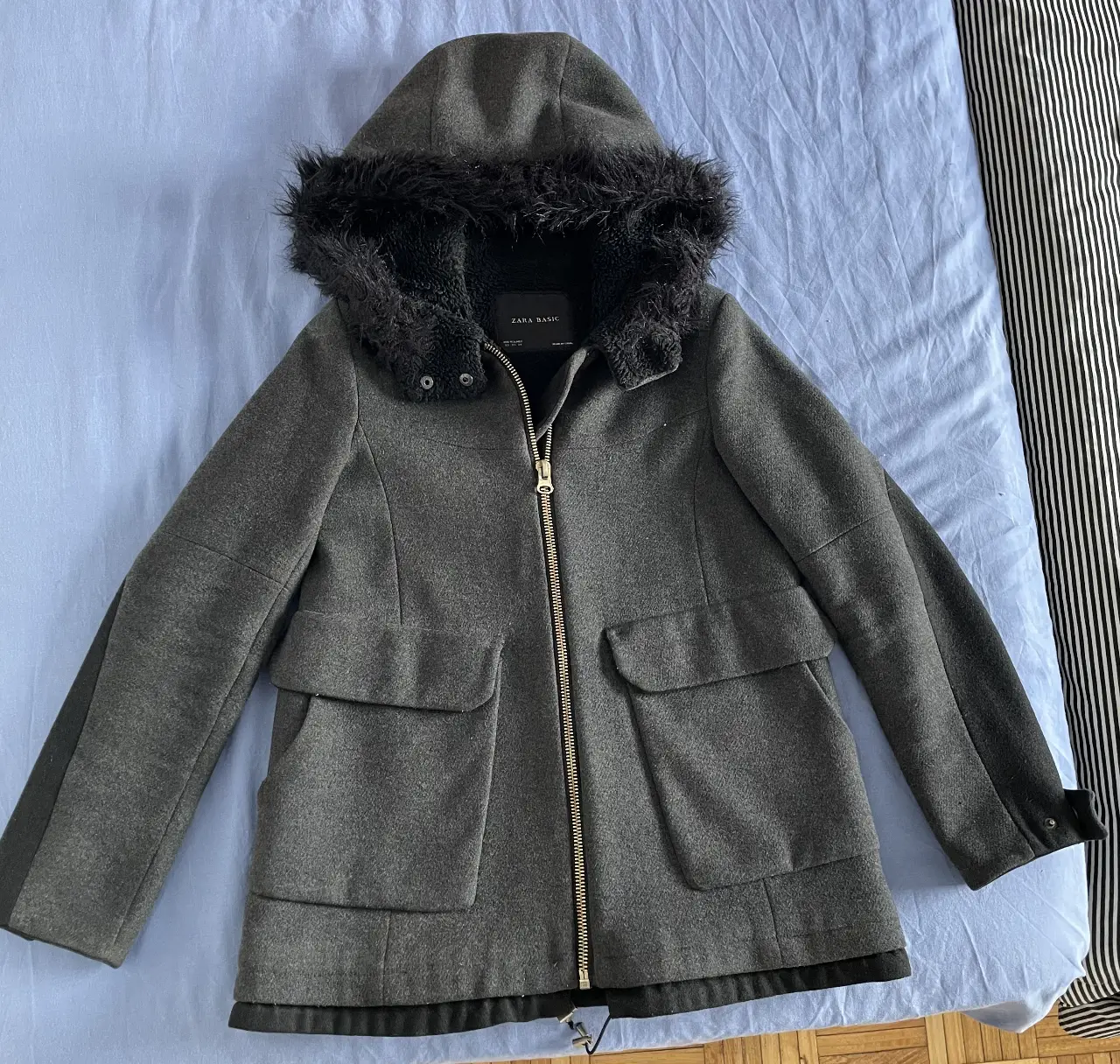 Tapado de paño gris oscuro con capucha y borde de piel sintética negra. Interior de polar, muy calentito. Ideal para el invierno, de la marca Zara. Tiene cierre frontal dorado y bolsillos laterales amplios. La capucha no es desmontable. 