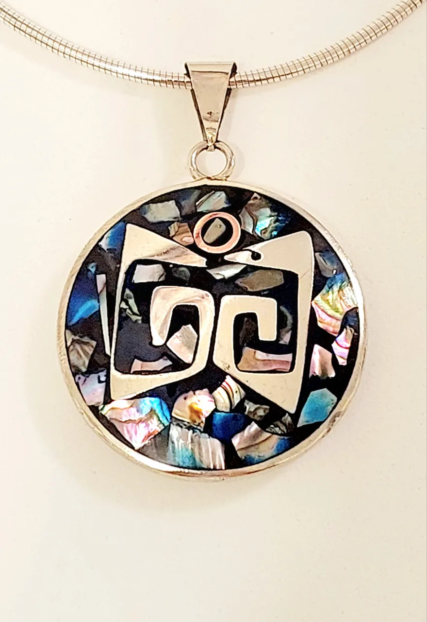 Este dije es una pieza de joyería artesanal mexicana (Taxco).
En el pasante tiene sello "Hecho en México" indica está fabricado en alpaca que a menudo se le llama "plata alemana" o "plata de alpaca", Concha Nácar y Abulón: Las incrustaciones iridiscentes (azules, verdes y rosadas) Estos materiales eran sagrados en la época prehispánica, asociados con las deidades del agua y la fertilidad.
La figura central es un glifo o símbolo de estilo prehispánico (Azteca o Maya). 
Glifo Escalonado: El diseño geométrico con ángulos rectos evoca la "Grecas" o el símbolo del Xicalcoliuhqui, que representa el ciclo de la vida, el movimiento de las nubes o la serpiente emplumada.La figura del dorso representa el árbol de la vida 
Diámetro dije: 4,5cm
