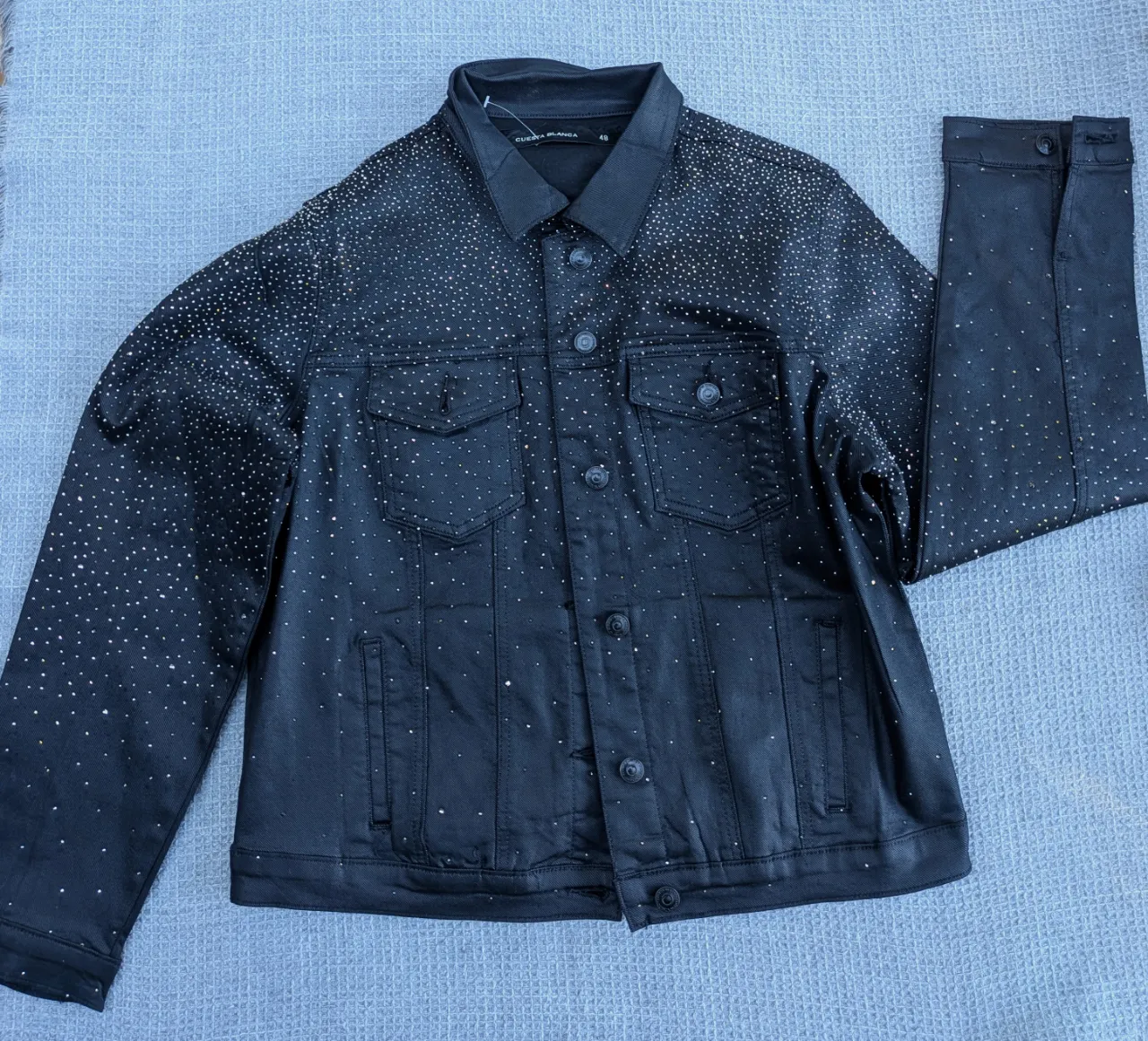 Campera de jean negra con brillo sutil tipo metalizado y tachas de brillos plateados en toda la prenda. Posee cuello camisero, botones al frente y bolsillos con tapa en el pecho.